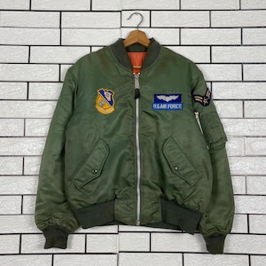 Könnte beinhalten: Eine olivgrüne Bomberjacke mit silbernem Frontreißverschluss, gerippten Bündchen und Saum sowie einem orangefarbenen Innenfutter. Auf der Brust befindet sich rechts ein gelb-blaues Emblem und links ein blauer Aufnäher mit weißen Flügeln und dem Text U.S. AIR FORCE.