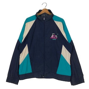 Op de afbeelding: Een vintage Adidas trainingsjack in marineblauw, teal en crème. De jas heeft een volledige ritssluiting, een hoge kraag en het woord "TEAM" met het Adidas-logo in roze en teal. De jas hangt aan een houten kleerhanger.