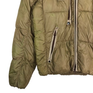 Puede incluir: Una chaqueta acolchada de color verde caqui con cierre de cremallera y un detalle de cremallera marr&oacute;n en el bolsillo izquierdo. La chaqueta tiene un dise&ntilde;o acolchado y un acabado ligeramente brillante.