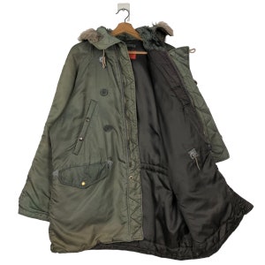 Op de afbeelding: Olijfgroene parka met een met bont gevoerde capuchon, een ritssluiting en knoopdetails. De jas heeft een gewatteerde binnenkant en meerdere zakken. De parka hangt aan een houten hanger.