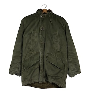 May include: An olive green parka jacket with a fur-lined hood, button fastenings, and large front pockets. The jacket is hanging on a metal hanger. The jacket appears to be made of a durable, heavy-duty material.