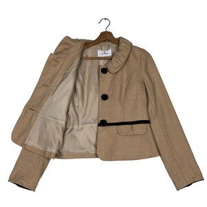 Op de afbeelding: Een beige blazer met zwarte details, waaronder drie knopen en een bies op de zakken. De jas is open en toont een lichtbeige voering. Hij hangt aan een houten hanger. De kraag is rond.