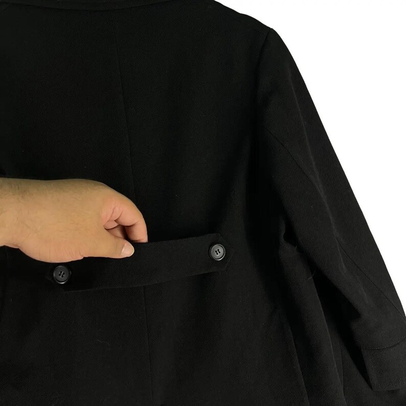 May include: A black coat with a buttoned pocket and two buttons visible. The coat is made of a smooth, dark fabric. The pocket is being held open by a hand.