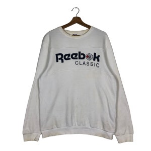 Op de afbeelding: Wit Reebok Classic sweatshirt met lange mouwen. Het sweatshirt heeft het Reebok-logo in marineblauw met een rood-witte afbeelding en het woord "CLASSIC" in het wit. Het sweatshirt hangt aan een houten hanger.