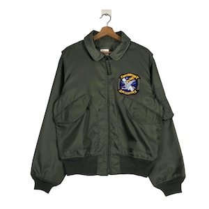May include: Olive green bomber jacket with a collared neckline, two front pockets, and a ribbed hem and cuffs. A patch on the chest displays a blue and yellow emblem with text. The jacket is hanging on a wooden hanger.