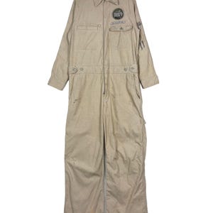 May include: A beige jumpsuit with long sleeves, a collared neckline, and a front zip. It has two chest pockets and patches with text and graphics. The jumpsuit is suspended from a wooden hanger.