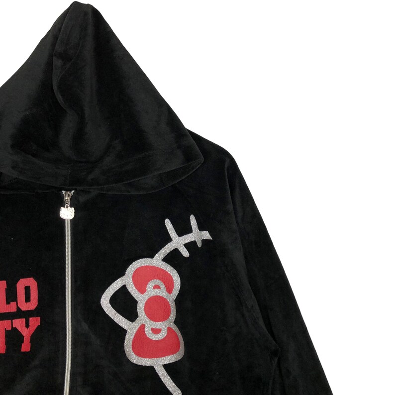 May include: Black velour hooded sweatshirt with a red and silver Hello Kitty graphic on the front. The graphic features Hello Kitty's signature bow and whiskers.