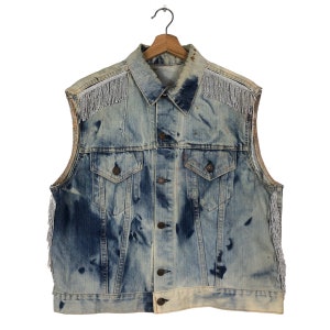 May include: A light blue denim waistcoat with silver fringe trim around the neckline and armholes. The waistcoat has a button-up front and two pockets.