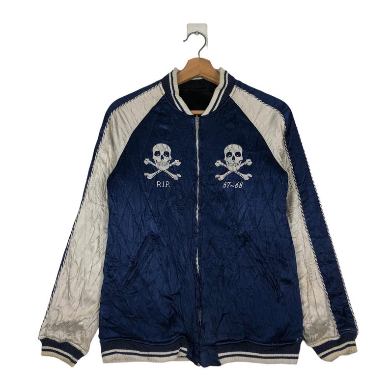 May include: A blue satin bomber jacket with white sleeves and a zipper closure. The jacket features two embroidered skull and crossbones designs, one on each side. The text "R.I.P." and "57-68" are embroidered below the skulls.
