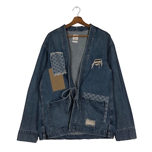 May include: A blue denim jacket with a tie closure and two front pockets. The jacket features decorative patches and distressed details. It is hanging on a wooden hanger.