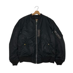May include: Black bomber jacket with a zip-up front, two front pockets, and a small pocket on the left sleeve. The jacket has a ribbed collar, cuffs, and hem. The jacket is hanging on a wooden hanger.