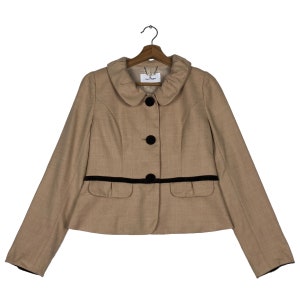 Op de afbeelding: Een beige jasje met een ronde kraag, zwarte knopen en een zwarte bies langs de taille. De jas heeft lange mouwen en twee kleine zakken. De jas hangt aan een houten kleerhanger.
