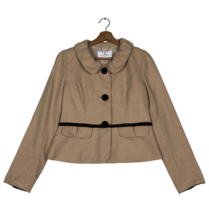 Op de afbeelding: Een beige jasje met een ronde kraag, zwarte knopen en een zwarte bies langs de taille. De jas heeft lange mouwen en twee kleine zakken. De jas hangt aan een houten kleerhanger.
