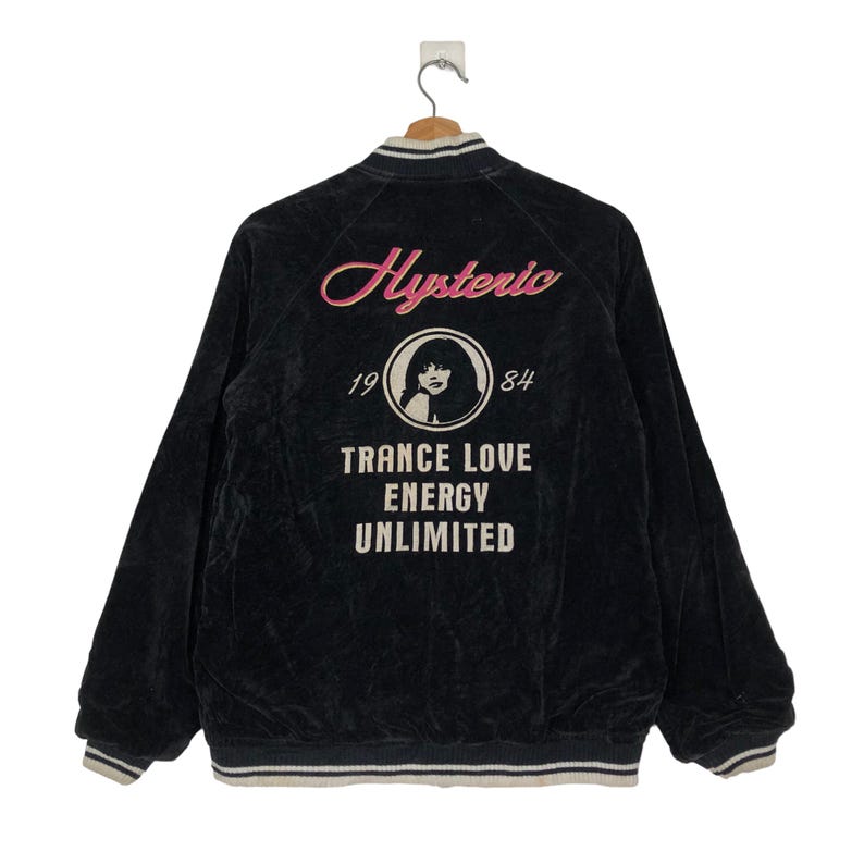 May include: Black velvet bomber jacket with a white and black striped collar and cuffs. The back of the jacket features a pink and white graphic with the text "Hysteric 19 84 Trance Love Energy Unlimited".