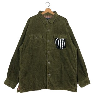 Op de afbeelding: Olijfgroen corduroy overhemd met een knoopsluiting, twee borstzakken en een skelet hand patch op de linkerzak. Het overhemd hangt aan een houten kleerhanger.