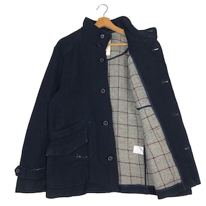 May include: A navy blue wool coat with a high collar and button closure. The coat features two front pockets and a plaid lining in shades of brown, red, and white. The coat is hanging on a wooden hanger.