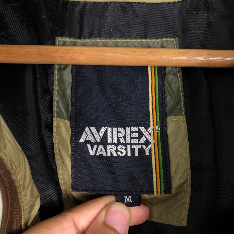 Puede incluir: Una etiqueta de chaqueta Avirex Varsity negra y verde con texto blanco y una etiqueta de talla M.