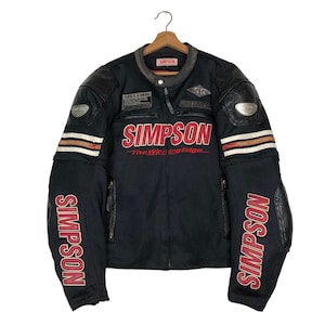 Op de afbeelding: Zwarte motorjas met de merknaam "SIMPSON" in grote rode letters over de borst. De jas heeft leren patches op de schouders en ellebogen, met witte en bruine strepen op de mouwen. De tekst "LIFE'S A DRAG" is zichtbaar.