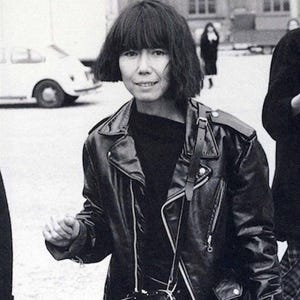 May include: Black and white photograph of a person wearing a black leather jacket with a zipper closure, a black top, and a black shoulder bag. The person has short, dark hair and is smiling. The background includes a building and a vehicle.