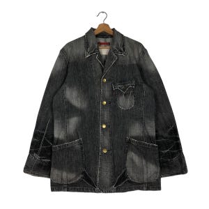 May include: A faded black denim jacket with a classic collar, four gold-colored buttons, and two large front pockets. The jacket has a small pocket on the chest and is hanging on a wooden hanger.