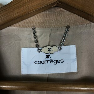 Op de afbeelding: Close-up van de binnenkant van een beige jas, met het merklabel "courr&egrave;ges". Het witte label heeft zwarte tekst en een logo, verbonden door een zilveren ketting. De jas hangt aan een houten kleerhanger.