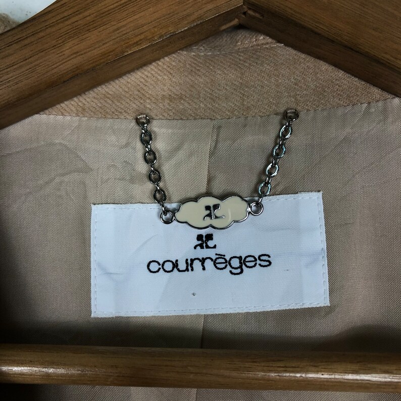 Op de afbeelding: Close-up van de binnenkant van een beige jas, met het merklabel "courr&egrave;ges". Het witte label heeft zwarte tekst en een logo, verbonden door een zilveren ketting. De jas hangt aan een houten kleerhanger.