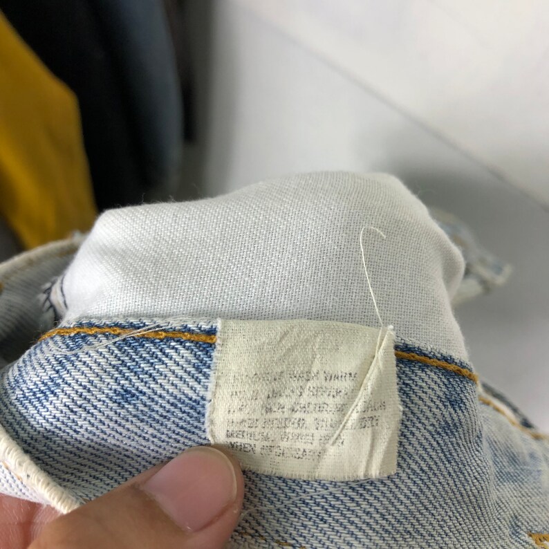 Może przedstawiać: Zbliżenie na niebieską metkę dżinsową z białym tekstem, kt&oacute;ry brzmi "Machine Wash Warm, Tumble Dry Low, Do Not Bleach, Iron Low, Do Not Dry Clean, Made in USA, 100% Cotton, When It's Jeans, It's Levi's."