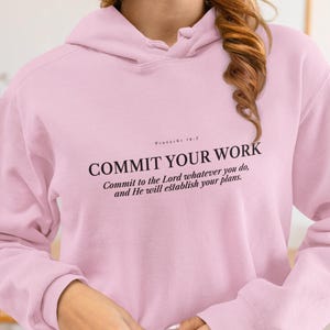 May include: A pale pink hoodie featuring the text "COMMIT YOUR WORK" in black, with a quote from Proverbs 16:3. The hoodie has a hood and long sleeves. The text reads: Commit to the Lord whatever you do, and He will establish your plans.