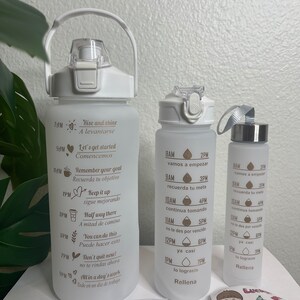 May include: Three white water bottles with motivational quotes and time markers. The bottles have gold lettering and are decorated with hearts and other symbols. The largest bottle has a handle. The smallest bottle has a silver lid. The bottles are on a white surface with a small bag of colorful stickers.