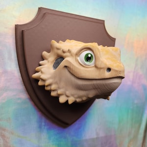 May include: A whimsical, tan-colored bearded dragon head sculpture with a friendly expression, mounted on a brown plaque. The lizard has a detailed, textured appearance, with a large green eye. The plaque is shield-shaped.
