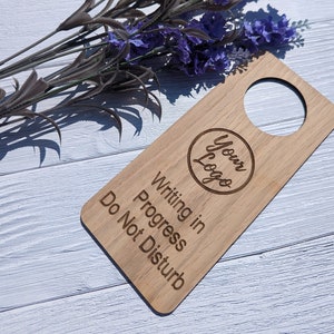 May include: Wooden door hanger with the text "Writing in Progress Do Not Disturb" and a circle with the text "Your Logo". The hanger is light brown and has a hole at the top for hanging on a door.
