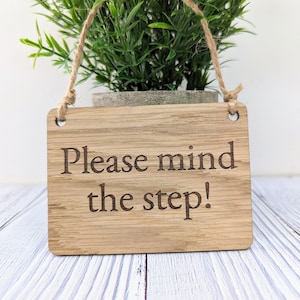 May include: A wooden sign with the text "Please mind the step!" hanging from a twine string. The sign is light brown and has a natural wood grain.