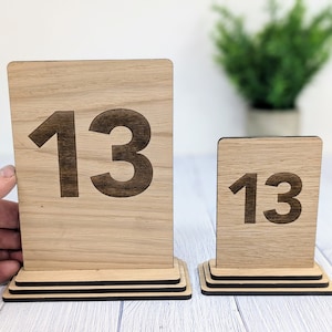 May include: Two wooden table number signs with the number 13 engraved on them. The larger sign is on a tiered stand, while the smaller sign is on a single stand.