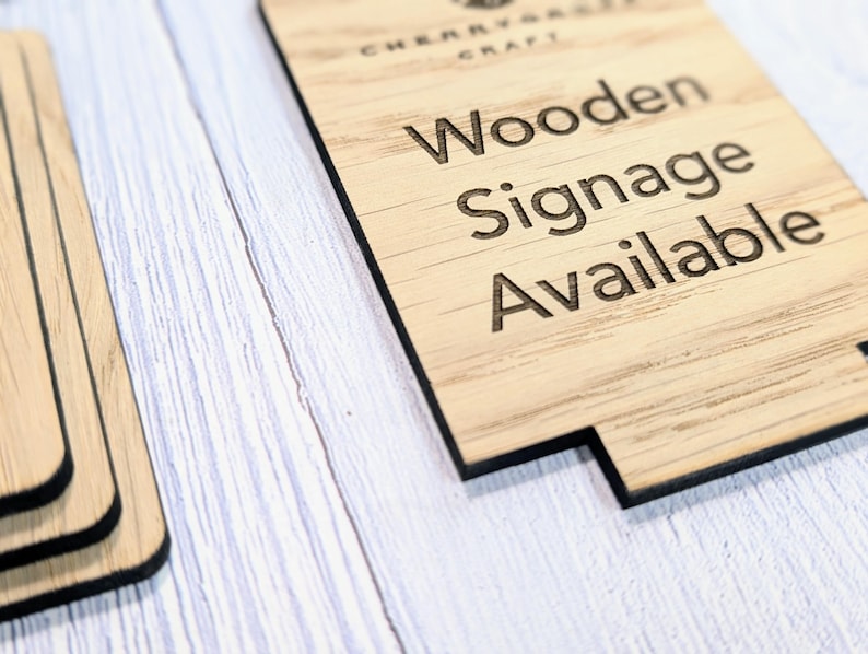May include: A close-up of a wooden sign with the text "Wooden Signage Available" engraved on it. The sign is made of light brown wood with a natural grain pattern.