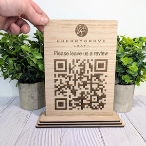 May include: A wooden sign featuring the text "CHERRYGROVE CRAFT" and "Please leave us a review" above a QR code. The sign is held by a hand and is displayed on a wooden surface. Two small potted plants are on either side of the sign.