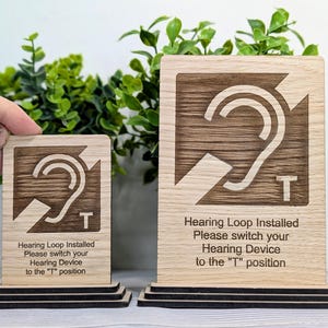 May include: Two wooden signs with a hearing loop symbol and the text "Hearing Loop Installed Please switch your Hearing Device to the "T" position". The signs are on a wooden base.