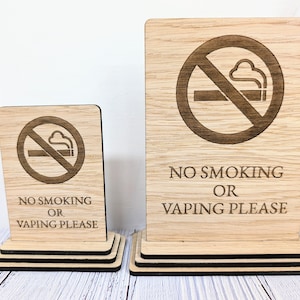 以下が含まれることがあります： 禁煙のシンボルと「No Smoking or Vaping Please」というテキストが書かれた木製の看板が2つ。看板は段になった木製の台座の上に置かれています。