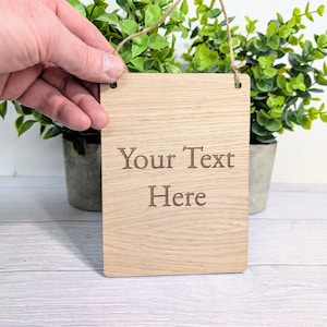 May include: A wooden sign with the text "Your Text Here" engraved on it. The sign is hanging from a string and is in front of two potted plants.