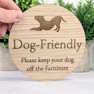 May include: A circular wooden sign with the words "Dog-Friendly" and "Please keep your dog off the furniture" engraved. A dog silhouette is above the text. The sign is held by a hand.