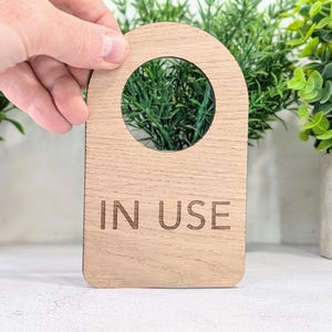 以下が含まれることがあります： 丸みを帯びた上部と円形の切り抜きがある、明るい色の木製ドアハンガー。「IN USE」の文字が木に刻まれています。ハンガーは手で持たれており、背景には緑が見えます。