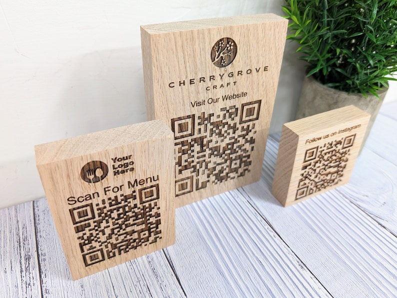 Op de afbeelding: Drie houten borden met QR-codes. Het bovenste bord zegt "Cherrygrove Craft Bezoek onze website" en heeft een QR-code. Het middelste bord zegt "Uw logo hier Scan voor het menu" en heeft een QR-code. Het onderste bord zegt "Volg ons op Instagram" en heeft een QR-code.