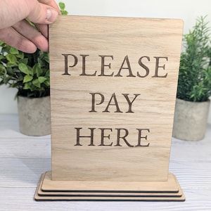 May include: Wooden sign with the text "PLEASE PAY HERE" in a rustic style. The sign is on a stand with three tiers.