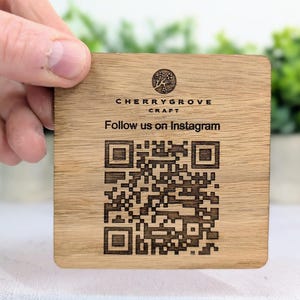 May include: A square wooden coaster with the text "CHERRYGROVE CRAFT" and "Follow us on Instagram". The coaster features a QR code and a tree logo. The coaster is held by a hand.