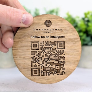 May include: A circular wooden coaster featuring the text "Cherrygrove Craft" and "Follow us on Instagram", plus a QR code. The coaster is held by a hand and has a natural wood grain finish.
