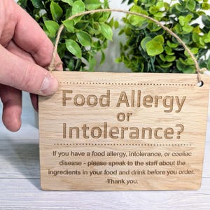 Könnte beinhalten: Ein Schild aus Holz mit dem Text "Food Allergy or Intolerance?" und einer Nachricht, die darauf hinweist, das Personal über Lebensmittelallergien oder -unverträglichkeiten zu informieren.