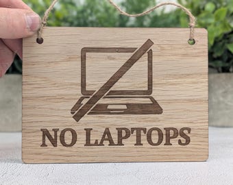 Op de afbeelding: Een rechthoekig lichtbruin houten bord met een donkerbruine lasergegraveerde afbeelding van een laptop met een diagonale lijn erdoorheen. De tekst NO LAPTOPS is in hoofdletters onder de afbeelding gegraveerd. Het bord heeft twee gaten met touw.
