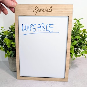 May include: A rectangular wooden framed whiteboard with the word "Specials" engraved at the top. The word "WIPEABLE" is written in blue marker on the white surface. Two small potted green plants are on either side of the whiteboard.