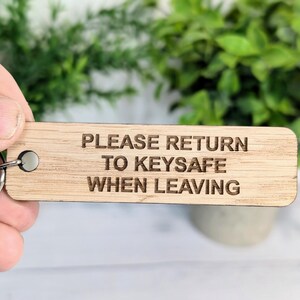May include: A wooden keyring with the text "PLEASE RETURN TO KEYSAFE WHEN LEAVING" engraved on it. The keyring is being held by a hand.