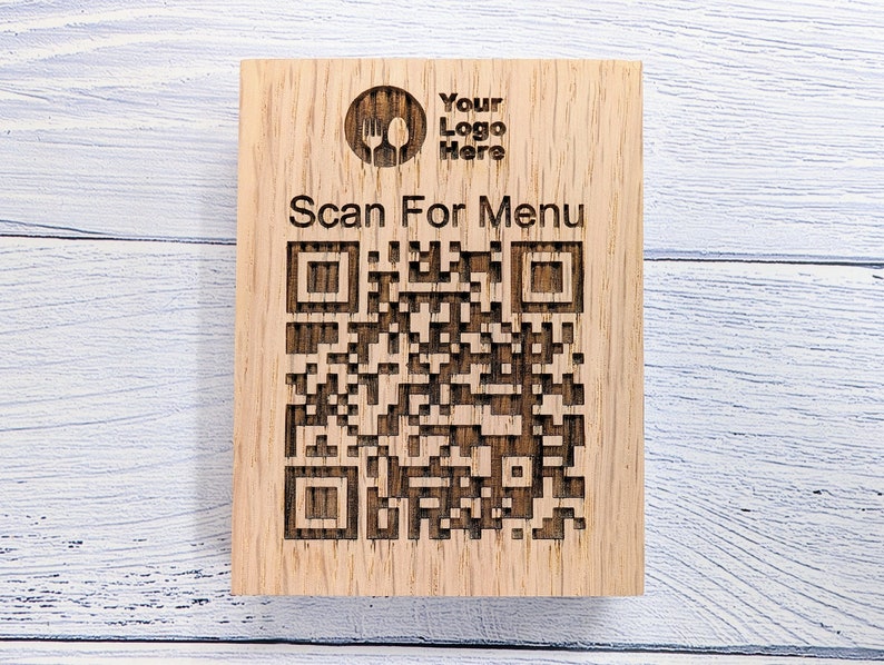 Op de afbeelding: Een houten bord met een QR-code waarop "Scan For Menu" staat. Het bord heeft ook een logo met een vork en een lepel en de tekst "Your Logo Here".