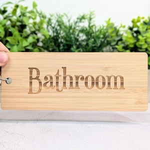 May include: A rectangular bamboo sign with the word "Bathroom" engraved in a serif font. The sign is attached to a silver key ring. The background features green plants.
